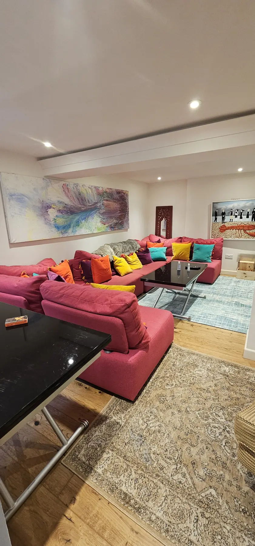 Large converted basement living room in Kent with a long red corner sofa, artwork, downlights and oak flooring